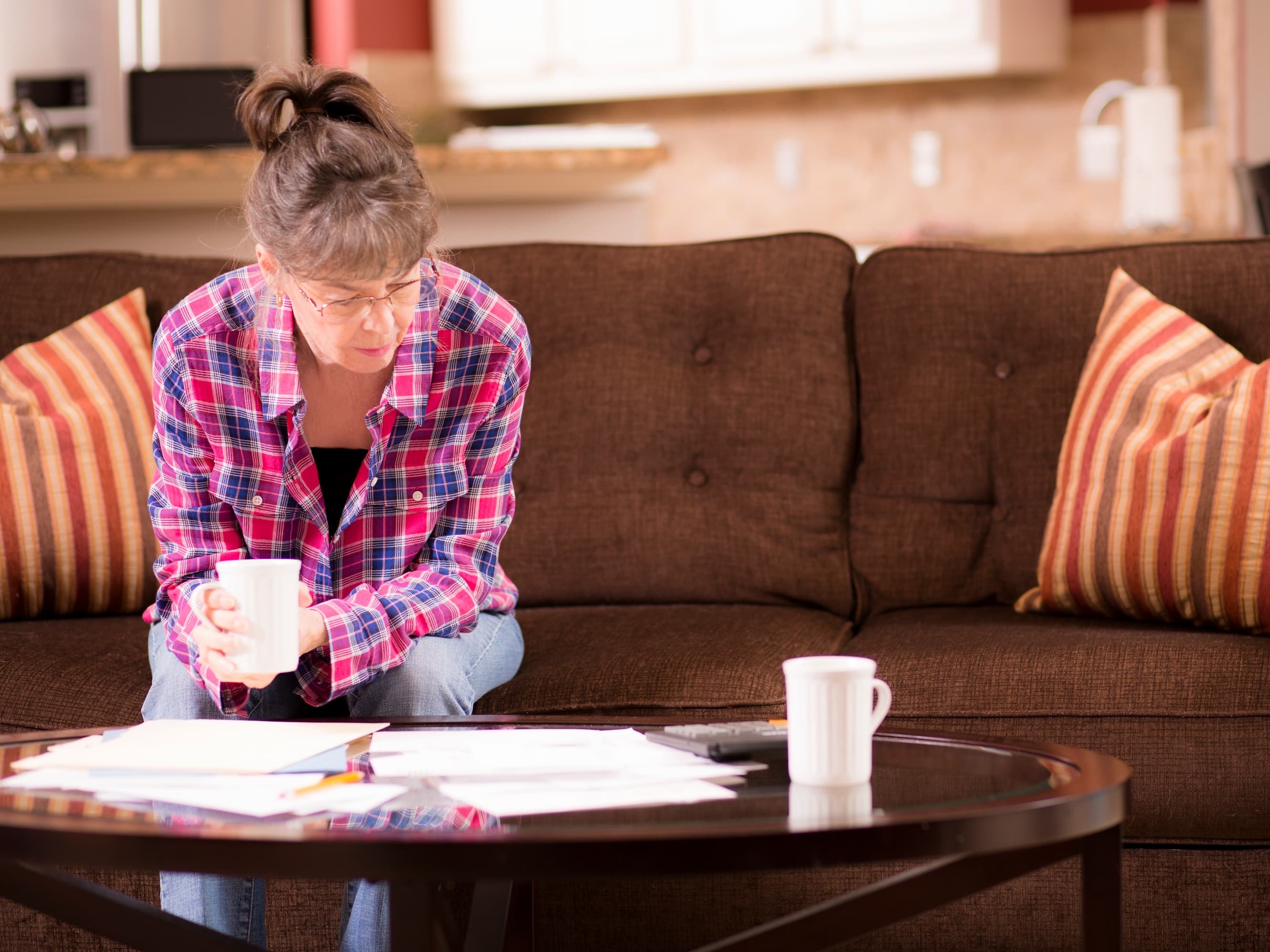 Woman sitting on a sofa, looking at paperwork on the table in front of her. She is holding a mug, and there is another mug on the table.