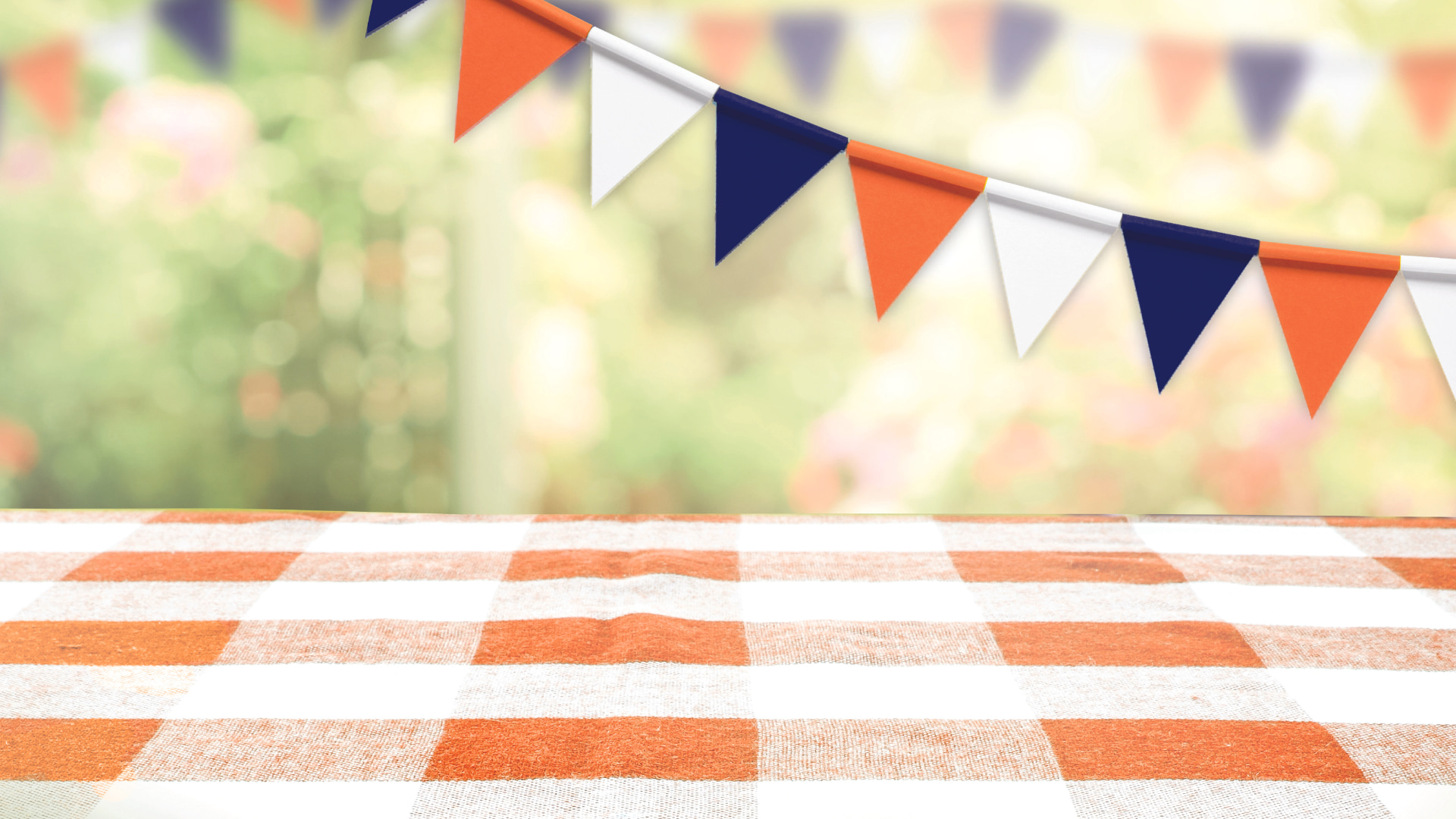 Orange and white checked tablecloth in the foreground, outdoors green blurry scene in the background, and bunting across the top right corner in orange, white and blue. Garden fete scene.