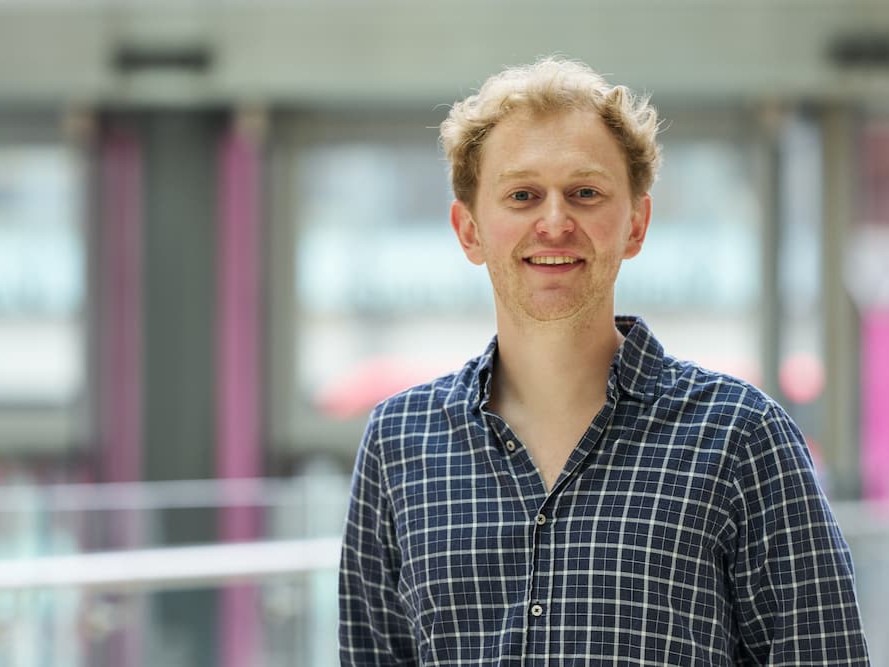 Dr Ben Clarke standing in a corridor at the Francis Crick Institute. He is smiling at the camera.