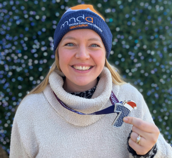 Lady smiling holding a 7 in 7 medal wearing a MNDA hat