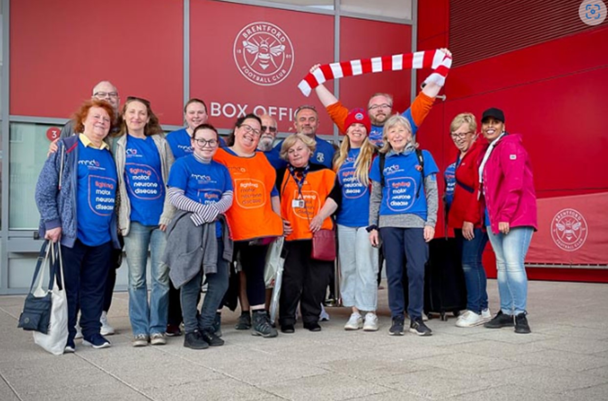 Branch volunteers in blue and orange MND Association t-shirts outsdide Brentford's stadium with one person holding a red and white scarf