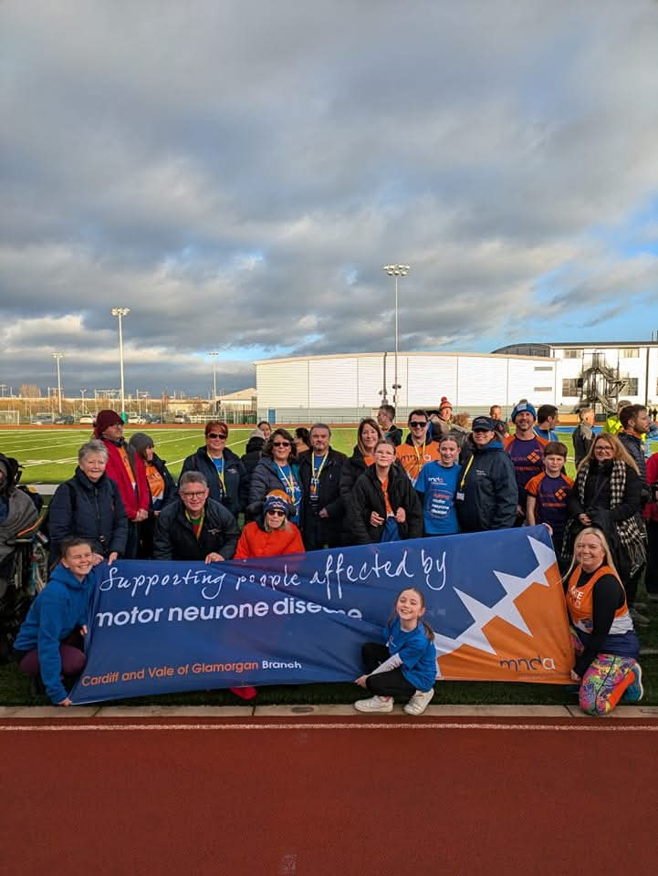 Two dozen supporters of the Cardiff and Vale Branch holding an MND banner at an athletics track on an overcast day