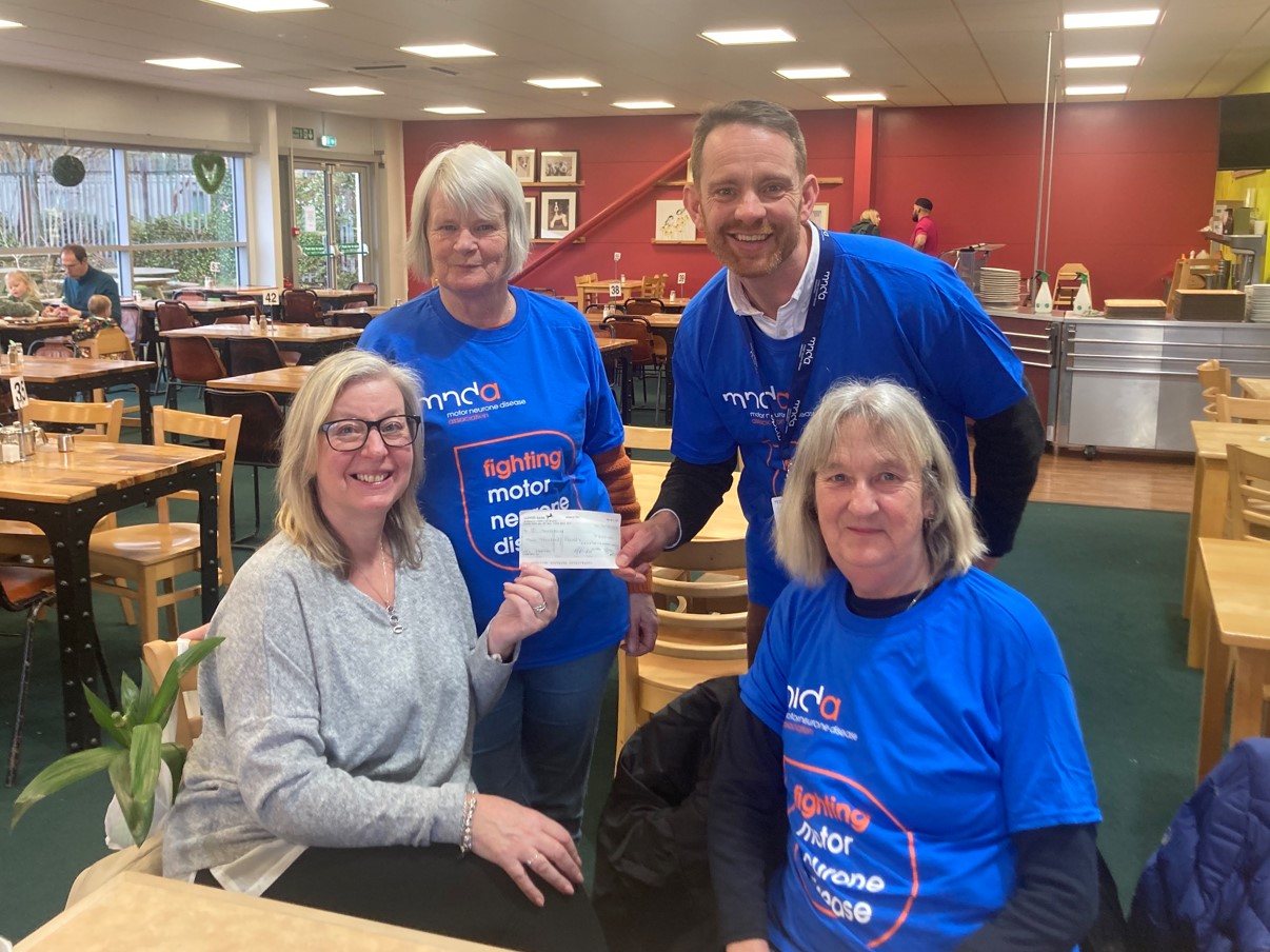 Janine a seated lady presenting a cheque to staff member Andy  in a blue MND Association t shirt also pictured are committee member lee and Caroline a person living with MND