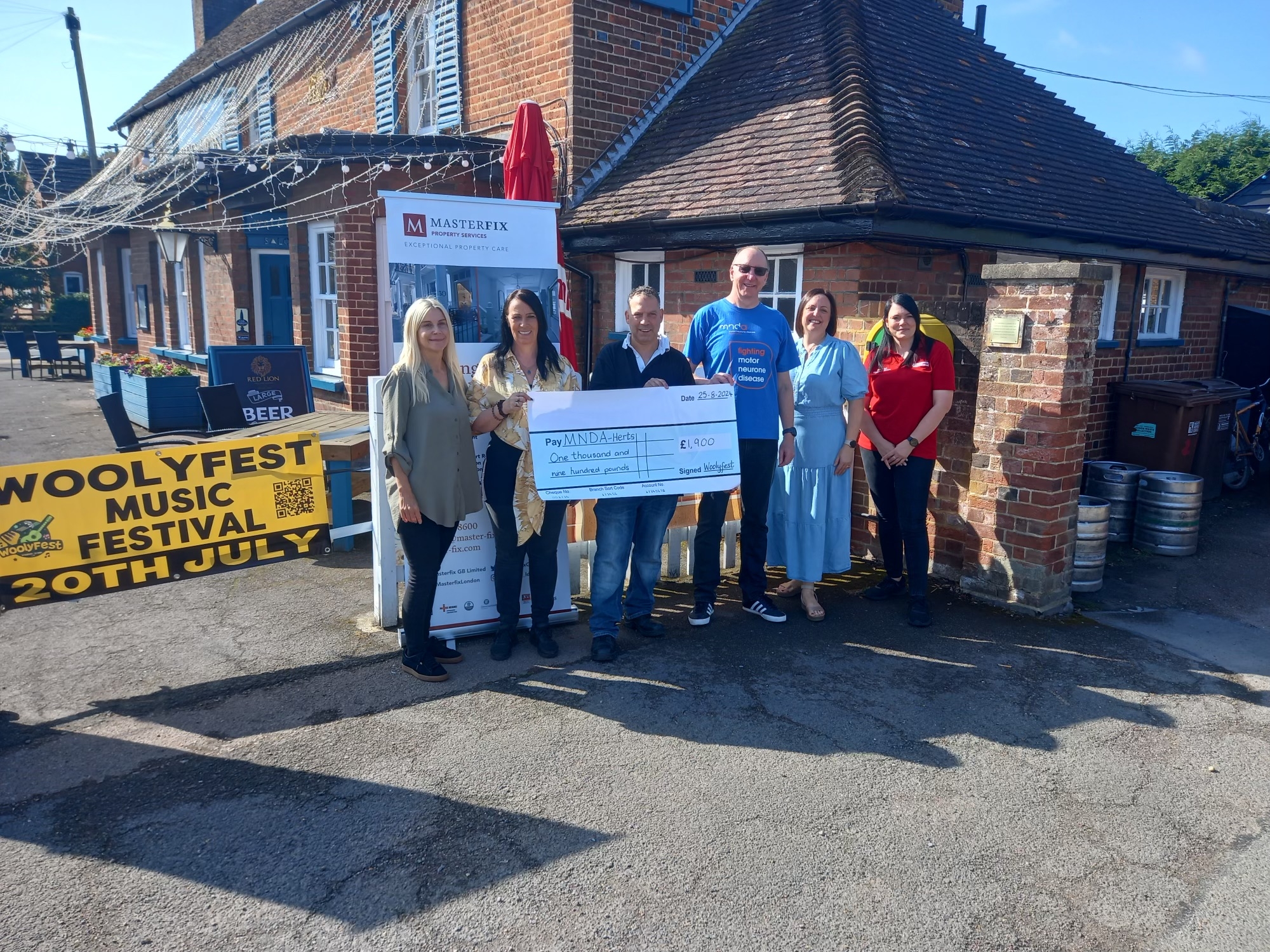 MNDA and Woolyfest volunteers stand presenting a cheque