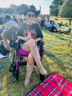 Debbie in her wheelchair with a can of Pimms enjoying an Ibiza Weekender festival. She sits with her legs crossed, smiling in a field. People are in the background enjoying the music. 